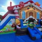 A colorful inflatable bounce house designed to look like a gingerbread house with candy decorations, a slide on the left side, and set up outdoors on a blue tarp under a sunny sky—perfect for party rentals Long Island celebrations.