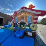 An inflatable bounce house Long Island, resembling a colorful candy-themed gingerbread house, stands on a blue tarp outdoors near a brick building and wooded area under the sunny NY sky—perfect for party rentals Long Island.