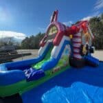 A colorful inflatable water slide with candy-themed decorations sits on a blue tarp in an outdoor lot, surrounded by trees and a partly cloudy sky—perfect for NY party rentals Long Island or any bounce house Long Island event.