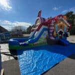A colorful inflatable bounce house Long Island, styled as a gingerbread house with a slide, is set up outdoors on a blue tarp under a mostly sunny sky. Trees and buildings are visible in the background.