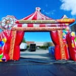 A large, colorful inflatable carnival entrance from party rentals Long Island features red and white stripes, balloons, and a Ferris wheel design. Set outdoors in NY under a blue sky, "Welcome to the Carnival" is written across the top.