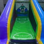 A colorful inflatable soccer game with a goal and an illustrated goalkeeper target—perfect for party rentals Long Island. With vibrant purple, blue, yellow, and green sides, this outdoor game setup adds fun to any NY celebration.