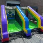 An inflatable dual-lane slide in vibrant purple, blue, yellow, and green with a soccer goalie graphic at the back—perfect for outdoor fun! Ideal for party rentals Long Island and bounce house Long Island events.