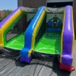An inflatable, dual-lane sports game from party rentals Long Island features two green lanes with blue, yellow, and purple borders, leading to a backdrop with soccer players and target circles. The game is set up outdoors on pavement.
