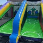 Inflatable soccer game setup with two lanes, each featuring a goalkeeper and target holes for scoring goals. Perfect for outdoor fun at party rentals Long Island, NY—set up near a brick building and garage door.