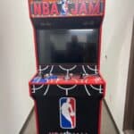 A vintage-style NBA Jam arcade game machine with red and black detailing, joystick, and buttons sits in a narrow hallway—perfect for party rentals Long Island, NY or as a unique addition alongside a bounce house Long Island event.