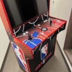 An NBA Jam-themed arcade machine with two control panels, joysticks, and NBA logos stands in a hallway on a carpeted floor—perfect for party rentals Long Island or adding excitement to any event in NY.