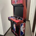 An NBA Jam arcade machine with a basketball-themed design, two joysticks, multiple buttons, and the NBA logo on the side stands in a hallway with gray carpet and white walls—perfect for NY party rentals Long Island events.
