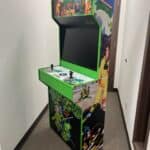 A Teenage Mutant Ninja Turtles arcade cabinet with colorful graphics stands in a narrow hallway—perfect for NY party rentals Long Island or adding retro fun to any bounce house Long Island event. It features two joysticks and several buttons on its control panel.