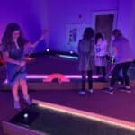 Four people play mini-golf indoors under purple lighting. One prepares to hit a ball while three others stand by different holes, holding clubs and focused on their shots—perfect for party rentals Long Island, NY.