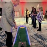 A man in a blazer prepares to hit a golf ball on an indoor mini-golf course, while four people watch. The room, set up for a party rentals Long Island event, features patterned carpet, bright lighting, and red accents.