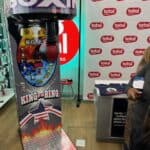 The Boxing Machine Arcade Game stands indoors near a display and counter with people nearby. Ideal for party rentals in Long Island, this machine features a punching bag and vibrant, colorful graphics.