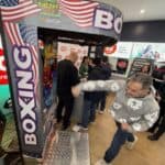 A man in a gray patterned hoodie punches the Boxing Machine Arcade Game with bold "BOXING" text and American flag decorations in a busy NY store, as several people watch and interact nearby.