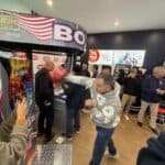 A man punches the Boxing Machine Arcade Game in a busy indoor arcade while others watch and wait. Some guests, likely from party rentals Long Island events, use their phones as various arcade machines and games line the walls.