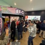 People gather in a lively arcade, some playing on the Boxing Machine Arcade Game decked with American flag decor. Others watch, chat, or snap photos—just like the upbeat energy at bounce house Long Island party rentals in NY.