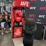 At a New York event, someone with long, dark hair punches the Boxing Machine Arcade Game as a crowd watches. The game displays a score and says "Test Your Knockout Power." Find Boxing Machine Arcade Game rentals on Long Island for your next party!.