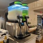 A commercial frozen drink machine on a countertop dispenses white and green slushies. Nearby are Corona Extra beer boxes—perfect for party rentals Long Island events—with assorted items and part of a person’s arm in the background.