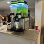 A dual frozen drink machine with two large clear containers holds green and white slushies on a countertop—perfect for party rentals Long Island. A woman stands behind the counter, and a red cup sits nearby.