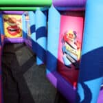 Colorful inflatable obstacle course with blue, purple, and green arches; bright posters and designs decorate the walls along a narrow, shaded pathway—perfect for party rentals Long Island, NY.