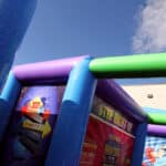 Colorful inflatable obstacle course under a blue sky with bright purple, green, and blue beams, plus a numbered game board on the left side. Perfect for party rentals Long Island, NY. Part of a building appears in the background.