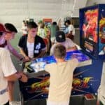 A group of young people gather around a Street Fighter II pinball machine, focused on playing, inside a brightly lit arcade—perfect for party rentals Long Island, NY, or fun events featuring a bounce house Long Island style.