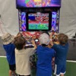 Four young children stand in front of a colorful Marvel-themed arcade machine, playing a retro video game together at a party rentals Long Island event. The arcade cabinet features comic book characters as the kids stay focused on the screen.