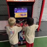 Two young boys in light-colored shirts play an NBA Jam arcade basketball game together at a NY gym, with the machine beside a chain-link fence—perfect for party rentals Long Island style.