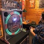 A person stands in front of an arcade game called "The Vault," featuring a large circular display with colorful lights and a digital timer, inside a wood-paneled restaurant—perfect for party rentals Long Island, NY.