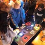 A group of young people gather around a Pac-Man Smash arcade game table, some pressing lit buttons while others watch and wait their turn in a cozy indoor setting—perfect for party rentals Long Island, NY.
