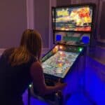 A woman with blonde hair, wearing a dark dress, plays a brightly lit pinball machine at a party rentals Long Island event in a dimly lit room with blue accent lighting. The pinball machine displays colorful graphics on its screen.