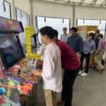 A group of young men, dressed in semi-formal shirts and pants, play on colorful arcade game machines at a NY party rentals Long Island event in a bright room with large windows. Some focus on the games, while others watch or wait their turn.