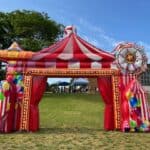 An inflatable red and white circus tent arch decorated with colorful balloons stands on a grassy field in NY, perfect for party rentals Long Island, with trees and a building in the background under a blue sky.