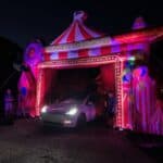 A white car is parked under a circus-themed inflatable archway at night, glowing with pink and purple lights. Perfect for party rentals Long Island, the arch is adorned with balloons and circus designs as two people stand on either side.
