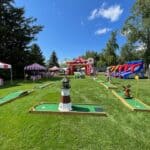 Outdoor carnival scene with red and white striped tents, bounce house Long Island inflatables, and mini-golf courses on green grass under a blue sky with scattered clouds. A small lighthouse decoration stands on one mini-golf course.