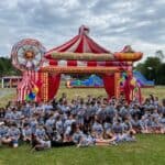 A large group of children in matching gray shirts gather in front of an inflatable red and white circus tent with colorful decorations and balloons at an outdoor field, featuring a bounce house Long Island obstacle course in the background.