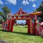 A large, colorful inflatable carnival entrance with red and white stripes, balloons, and a "Welcome to the Carnival" sign stands on a grassy field under a blue sky—perfect for party rentals Long Island or your next bounce house Long Island event.