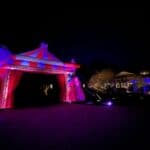A circus-themed entrance structure, brightly lit with red and purple lights, stands outdoors at night—perfect for party rentals Long Island, NY. Nearby trees and a house are illuminated with colorful accent lights in the darkness.