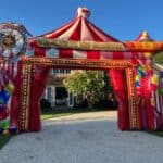 A large, colorful inflatable archway from party rentals Long Island, decorated with circus-themed designs and balloons, stands over a driveway with a "Welcome to the Carnival" sign. A house and trees are visible in the NY background.