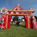 An inflatable carnival entrance from party rentals Long Island, decorated with stripes, balloons, and a Ferris wheel design, stands on grass. People gather beyond the archway under a blue NY sky at an outdoor event.