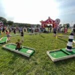 People play mini golf on a grassy field at an outdoor event with a red and white striped tent, bounce house Long Island, a large spinning wheel, and carnival decorations in the background. Families and children are enjoying the sunny day.