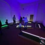 People, including children and adults, play mini-golf indoors on several glowing courses under dim, colorful lighting in a large room with high walls—perfect for NY party rentals Long Island or fun gatherings.