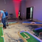 Several people in NY are playing mini golf indoors on a carpeted floor, with wooden courses set up and glowing blue LED lights—perfect for party rentals Long Island style. Some players are putting while others watch and cheer.