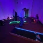 People play mini-golf on glow-in-the-dark courses in a dim indoor space, illuminated by neon lights in blue, green, pink, and yellow. Several adults and children enjoy the game—a fun option for party rentals Long Island, NY.