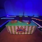 A mini-golf setup indoors with glowing course edges, a green-striped table displaying golf clubs and a basket, all under blue and purple lighting—perfect for party rentals Long Island, NY alongside your bounce house Long Island favorites.