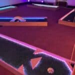 A brightly lit indoor mini-golf course in NY features colorful LED borders, artificial turf, obstacles like a small bridge and windmill—all set on a carpeted floor. Perfect for party rentals Long Island looking to add unique fun to any event.
