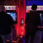 Two men play on side-by-side NBA Jam arcade machines in a dimly lit room with red and blue lighting at a party rentals Long Island event. Another person stands nearby, partially visible on the left.