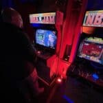 A person stands in front of a lit-up NBA Jam arcade machine in a dimly lit room, with another game next to it—perfect inspiration for party rentals Long Island, bringing classic arcade vibes to your next NY event. Red and blue lights set the mood.