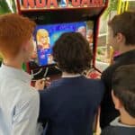 Three boys in suits stand closely together, playing a classic NBA Jam arcade game at a party rentals Long Island event. The arcade machine displays colorful graphics as the boys stay focused on the screen and controls.