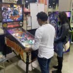 Two people stand at a Maverick-themed pinball machine in a large indoor event space, surrounded by arcade machines and booths—perfect inspiration for party rentals Long Island, NY, or pairing with a bounce house Long Island for your next event.