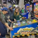 A group of people gathers around a blue "Road Show" pinball machine at an indoor arcade event in NY. The busy area features other arcade games and attendees, perfect for those seeking party rentals Long Island loves.